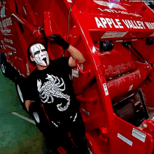 a man in a black shirt with a scorpion on it stands in front of an apple valley trash truck