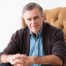 a man wearing a blue shirt and a black sweater is sitting in a chair with his hands folded