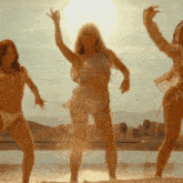 three women in bikinis are dancing on a beach in front of the sun