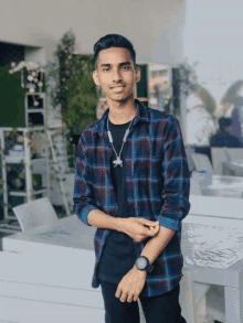 a young man wearing a plaid shirt and a watch stands in front of a table