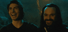 a man with a beard is smiling next to another man with long hair