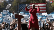 a woman in a red coat is standing at a podium in front of a crowd holding a sign that says bernie