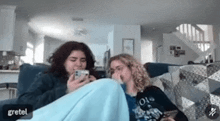 two women are sitting on a couch looking at their cell phones .