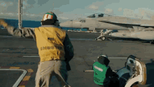 a man in a yellow vest with the word shooter on it is standing on a runway in front of a fighter jet .