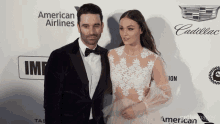 a man in a tuxedo and a woman in a white dress pose on a red carpet in front of an american airlines logo