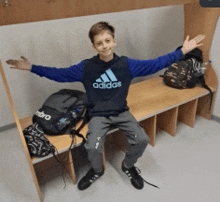 a boy wearing a blue adidas sweatshirt sits on a bench with his arms outstretched