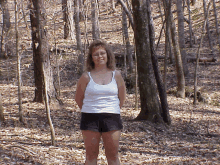 a woman in a white tank top and black shorts stands in a forest