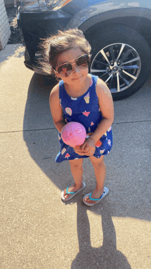 a little girl wearing sunglasses is holding a pink ball in front of a car