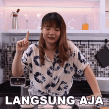 a woman in a floral shirt is standing in a kitchen with a sign that says " langsung aja "