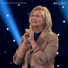 a woman in a tan jacket stands on a stage in front of a blue background with the words family feud canada on it