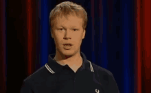 a man wearing a black shirt with a horseshoe on the front is standing in front of a red curtain .