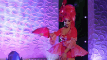 a drag queen in a pink dress is holding two pink fans