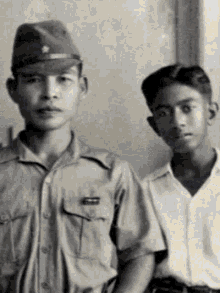 a black and white photo of two young men standing next to each other . one of the men is wearing a military uniform .