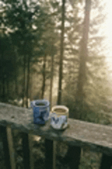 two cups of coffee are sitting on a wooden railing overlooking a forest .