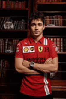 a young man in a red shirt is standing in front of a bookshelf with his arms crossed .