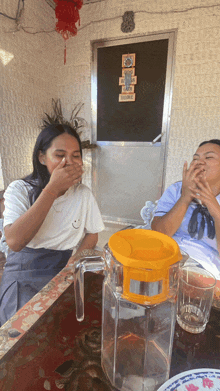 two women sit at a table with one covering her mouth