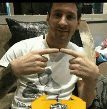 a man is sitting on a couch making a heart shape with his hands while wearing a white t-shirt .