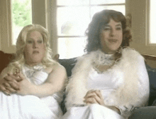 two women are sitting on a couch wearing white dresses