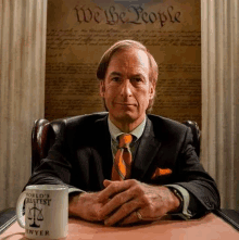 a man in a suit and tie is sitting at a table with his hands folded in front of a poster .