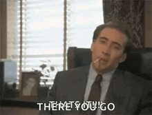 a man in a suit and tie is sitting in a chair smoking a cigarette and saying `` there you go '' .