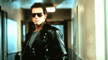 a man wearing sunglasses and a black leather jacket stands in a hallway