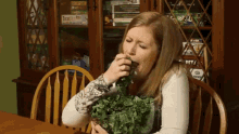 a woman sitting at a table with a bowl of greens in front of a cabinet with a board game called twister