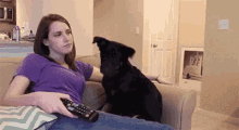 a woman is sitting on a couch holding a remote control and a dog is standing on her lap .