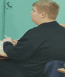 a boy in a black sweater is sitting at a desk with a pen in his hand