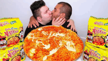 two men kissing in front of a large pizza made of ramen