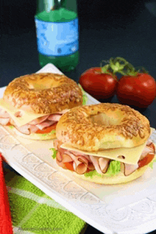 two bagel sandwiches on a white plate with tomatoes and a bottle of water in the background