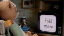 a teddy bear is sitting on a couch watching a television with the words esti mese on the screen .