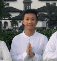 a young man in a white shirt is standing in front of a building with his hands folded in prayer .