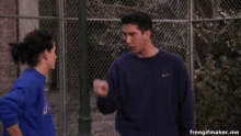 a man and a woman are standing next to each other in front of a chain link fence and talking .