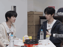 two young men are sitting at a table with bottles of water and a pot that has a sticker on it that says cup
