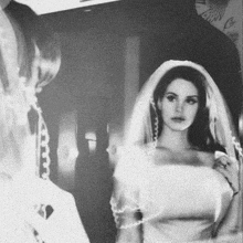 a black and white photo of a woman in a wedding dress and veil looking at herself in the mirror .