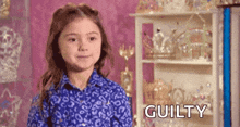 a little girl in a blue shirt is standing in front of a shelf with trophies and the word guilty .