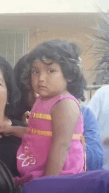 a little girl in a pink dress is looking at the camera while sitting in a chair .