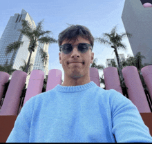 a man wearing sunglasses and a blue sweater takes a selfie in front of pink pipes