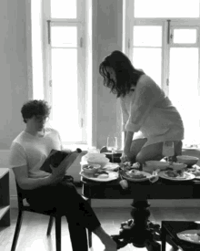 a man sits in a chair reading a book while a woman sits at a table with plates of food