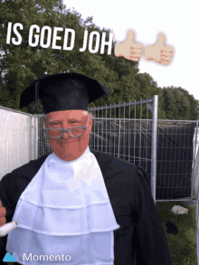 a man in a graduation cap and gown is standing in front of a fence with the words is goed joh above him