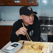 a man wearing a black hat is eating food with chopsticks in front of a container that says ' food ' on it