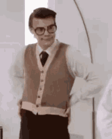 a man wearing glasses and a vest is standing in a room with his hands in his pockets .