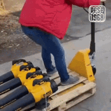 a man in a red jacket is pushing a yellow pallet truck with a bunch of blower heads on it .