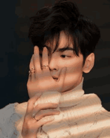 a young man wearing a turtleneck sweater and a ring on his finger is covering his face with his hand .