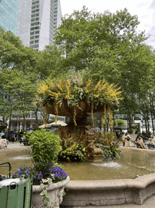 a fountain surrounded by flowers and a green trash can that says ' n.y.c.p. ' on it