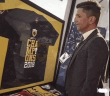 a man in a suit stands in front of a framed jersey that says champions 2018