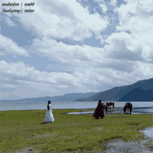 a woman in a white dress stands in a grassy field near a body of water with mountains in the background