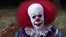 a clown with red hair and a red nose is standing in the grass .
