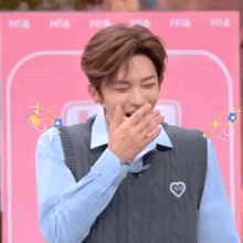 a young man is covering his mouth with his hand while wearing a blue shirt and a sweater vest .