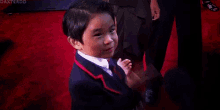 a young boy in a suit and tie is standing on a red carpet and making a funny face .